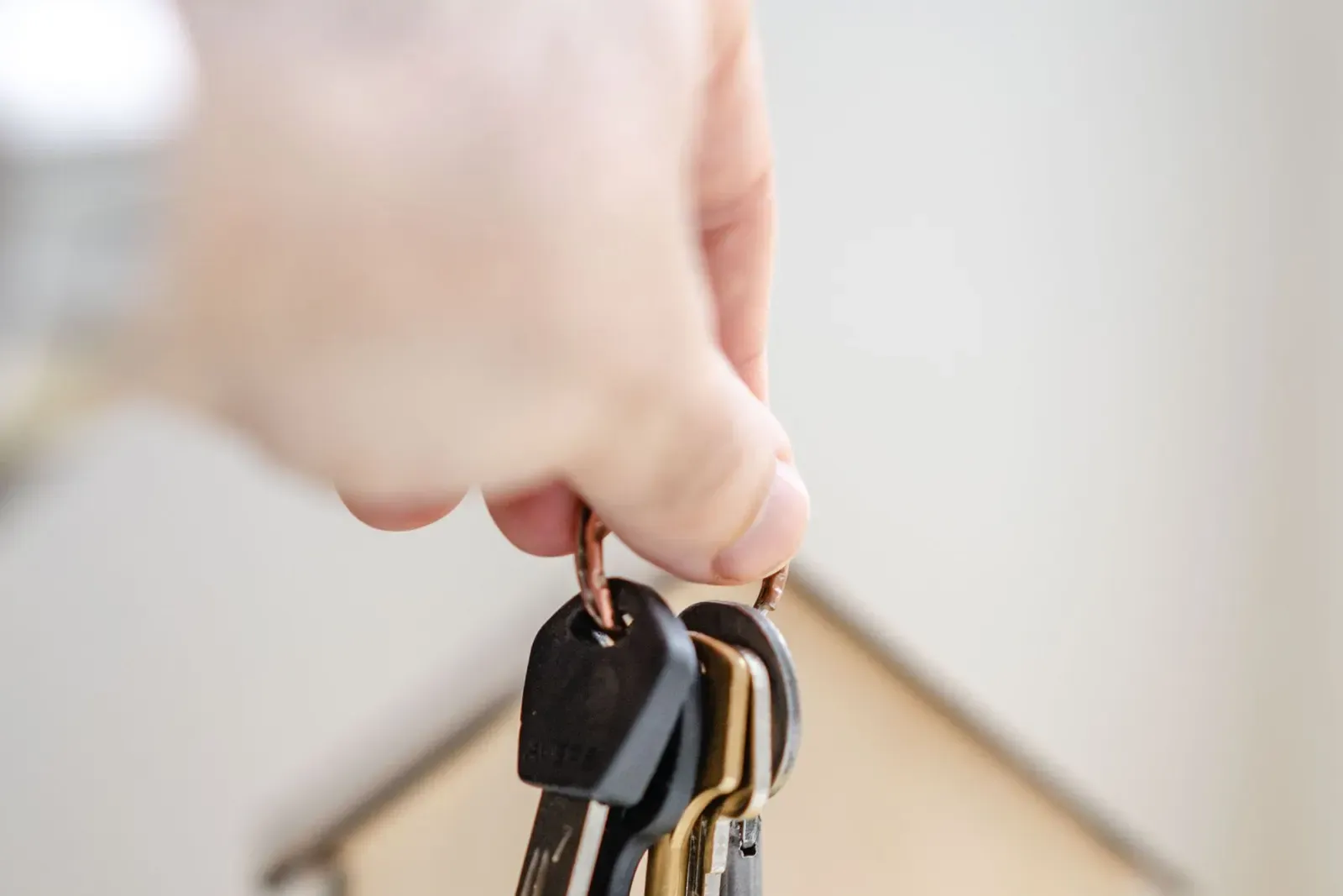 Hand holding a set of house keys in front of a small wooden house model