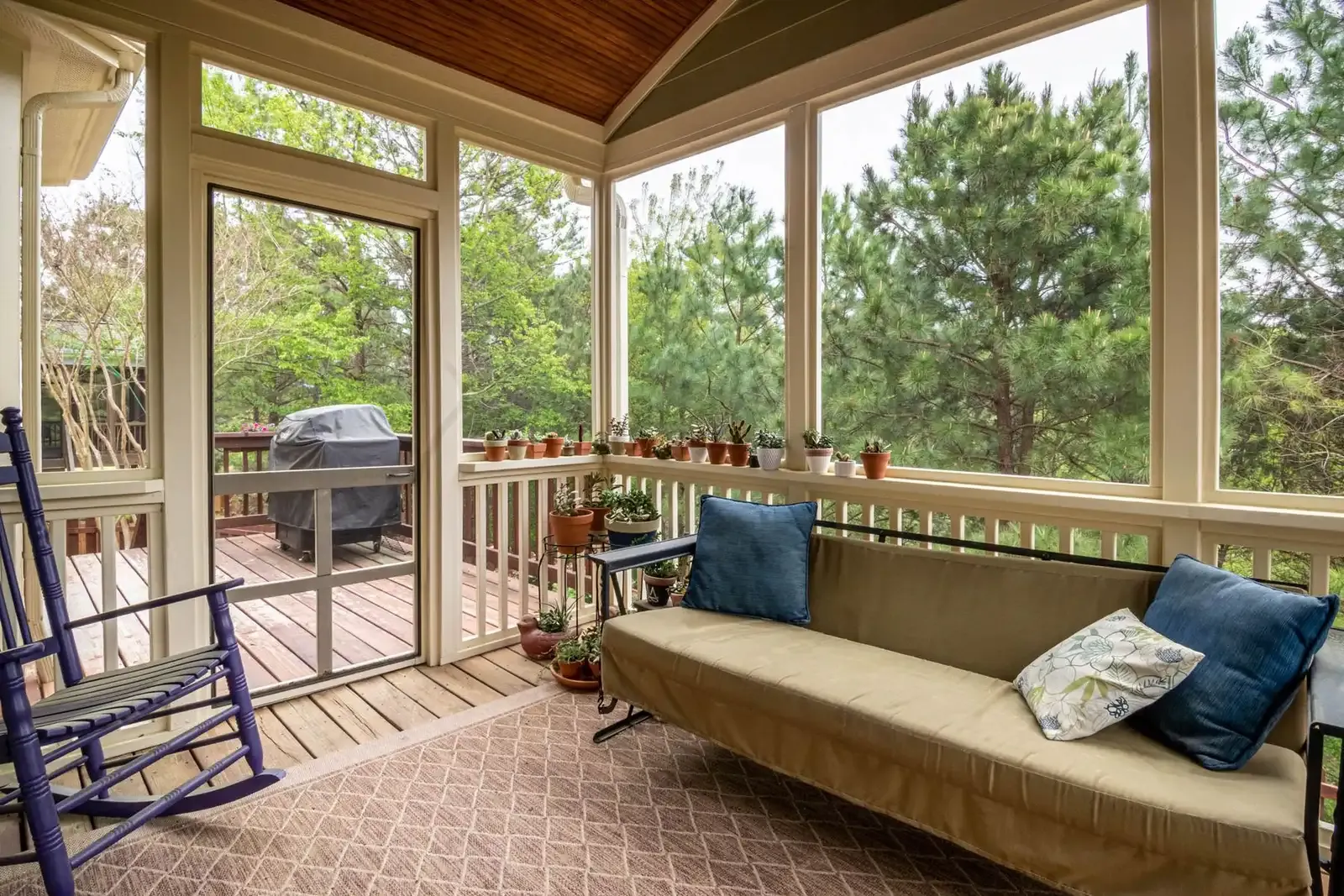Bright screened porch with a sofa and rocking chair overlooking green trees